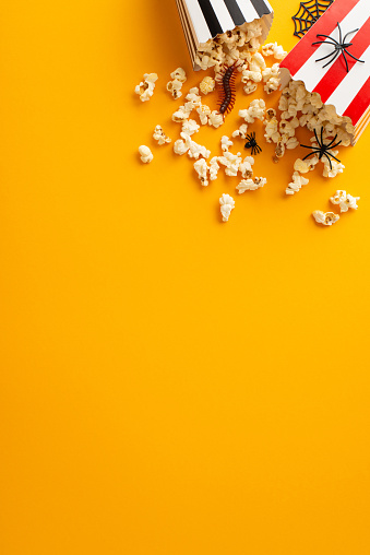 Experience spine-chilling horror on Halloween night. Above vertical view shows popcorn with spooky Halloween-themed decorations on an isolated orange background. Copyspace available for ads or text