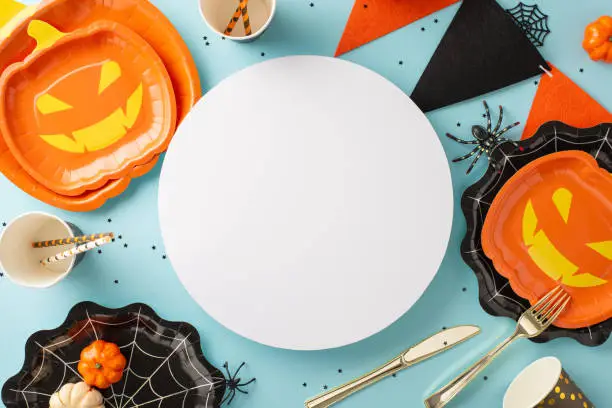 Unveil Halloween magic: Top view jack-o'-lantern and cobweb plates, cutlery, disposable cups, pumpkins, spiders, garland. Soft blue backdrop with circle offers prime real estate for your text or ad