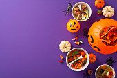 Embrace the wonder of kids' Halloween candy hunt. High-angle photo of a pumpkin baskets filled with candies and Halloween decorations on purple isolated backdrop, perfect for text or ad placement