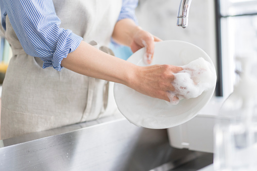 A Japanese woman with short hair is washing dishes while wearing an apron. Washed with dish detergent.Bright and clean kitchen with large wind
