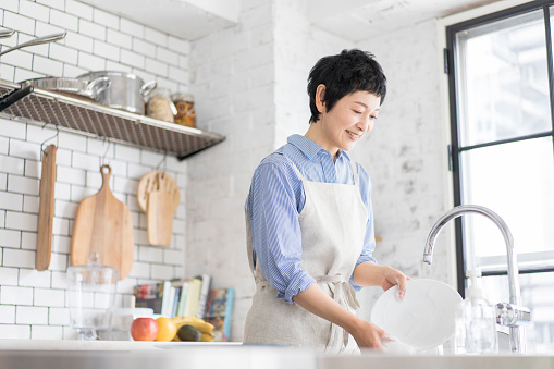 A Japanese woman with short hair is washing dishes while wearing an apron. Washed with dish detergent.Bright and clean kitchen with large windows.
