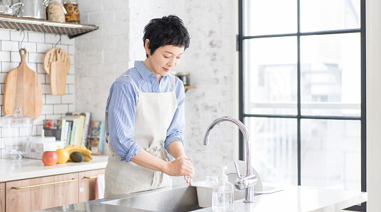 An Asian woman with short hair wearing an apron is washing her hands in the kitchen.Bright and clean kitchen with large windows.