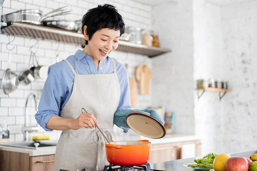 A Japanese woman is cooking, using a pot in the kitchen.She has a pot lid and a ladle.Stirring the soup.