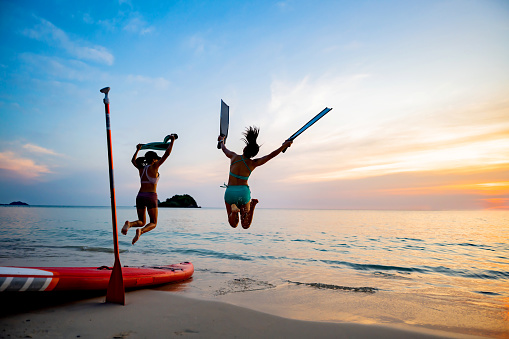 Asian athletic woman with paddle board with friend on beach sunset. Travel on summer holiday Thailand.