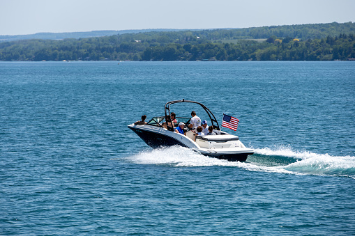 Group of friends driving n a speed boat. Taken on Skaneateles Lake in NY State.  Taken on a sunny day with calm lake water.  Shoreline in the background.