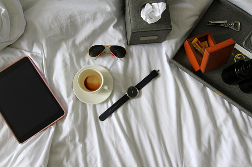 Top view of tablet, watch, black glasses, cup of coffee or tea, gray box of napkin, bottle opener, wooden box and plastic conditioner shampoo and bath shower gel on white bed. Working, relax and drink