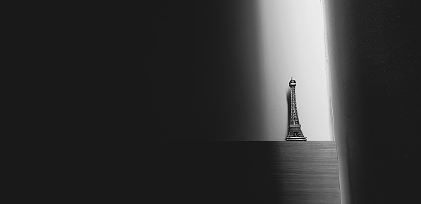 Small Eiffel tower put on wooden shelf on concrete wall background with sunlight from beside with copy space in black and white tone. Art of object with light in monochrome style.