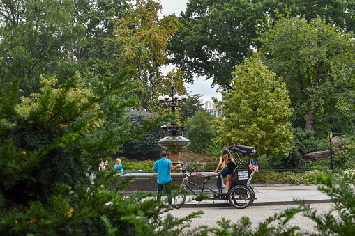 United States of America, New York City (NYC) - 2 people on a bike tour in Central Park front of a fountain. In the background, there are many trees and few tourists.