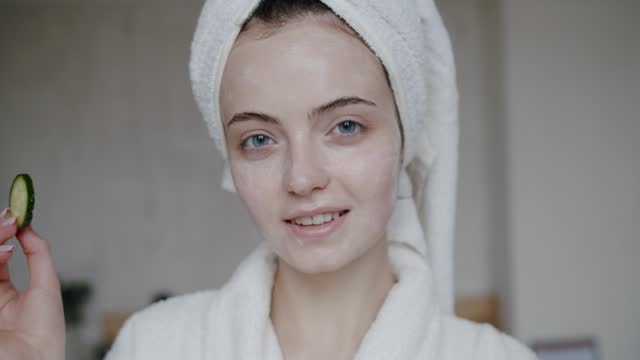 young woman in a bathrobe with a towel on her head covering eyes with cucumbers