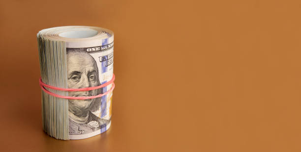 istock Many hundred dollar bills rolled up with a red rubber band on a colored background. 1669835140
