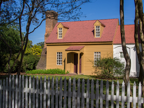 In Colonial Williamsburg, Virginia, a delightful two-story home features vivid yellow sidings, a bold red roof, and a prominent brick chimney. Against a blue sky, the house stands behind a white fence, radiating classic colonial charm.