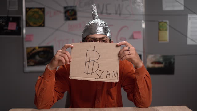 Male conspiracy theorist in a protective foil cap and glasses debunks myths. Conceptual cryptocurrency bitcoin with the word Scam
