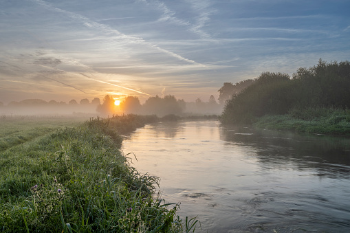 A gentle river scene with steady flow and grassy riverbanks featuring mist and the sunrising on the horizon