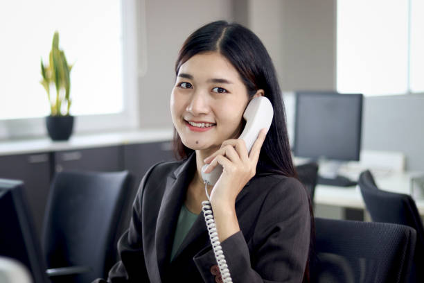 istock Beautiful Asian woman operator officer talking with customer by using landline phone at working desk in office with busy working colleagues blurred background. Smiling female receptionist at workplace 1669902821