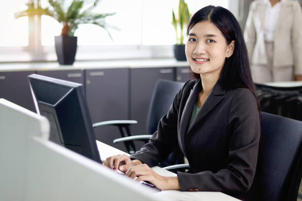 istock Happy beautiful Asian woman officer typing on desktop computer while sitting at office desk with blurred background of her busy working colleagues. Smiling female worker working at workplace. 1669904429