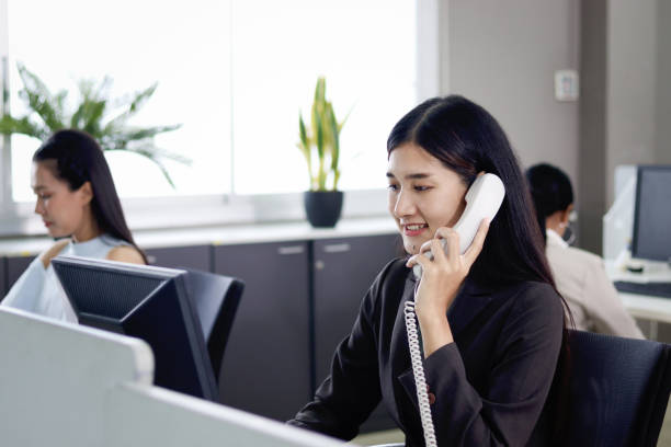 istock Beautiful Asian woman operator officer talking with customer by using landline phone at working desk in office with busy working colleagues blurred background. Smiling female receptionist at workplace 1669905564
