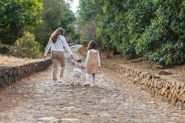Group of a mother walking outdoors with her two children holding hands along a scenic forest path enjoying nature on a sunny day.