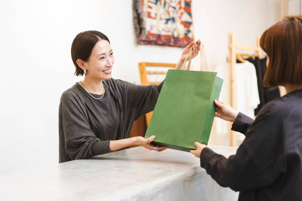 A Japanese woman works at a clothing store.She is serving customers. She is handing over the products purchased by the customer.