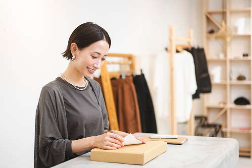 A Japanese woman works at a clothing store.She is putting the goods in the box.She is preparing the delivery.