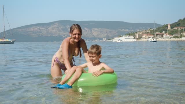 Happy mother in bikini helping her son seating in inflatable swimming ring and riding him in calm sea waves next to the beach.