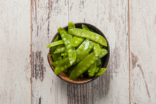 Top view of steamed fresh snap peas or mangetout in a hand made ceramic bowl on white weathered wooden table
