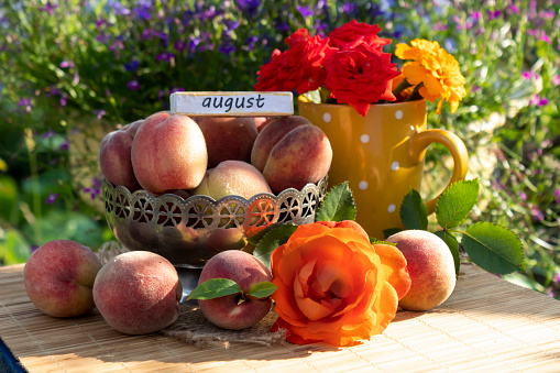 Against a background of bright greenery and flowers, there is a vase with ripe peaches, a bouquet of flowers, an orange rose and peaches.