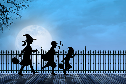 Three children scurry across a wood plank sidewalk lined with a wrought iron fence as they are silhouetted in front of a large rising moon on Halloween night.