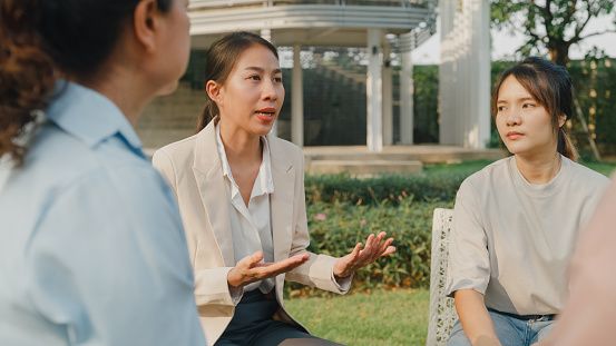 Group therapy session with diverse people sharing their sad problems and stories in park. Asian people sitting in circle at group therapy session with psychologist. Mental health concept.