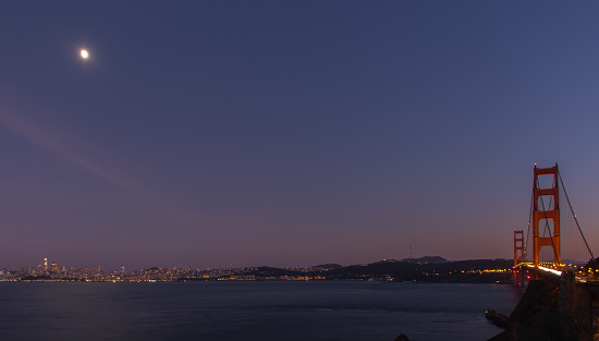Bridge illuminated under the moon at dusk, connecting San Francisco with beautiful nighttime skyline across the bay.