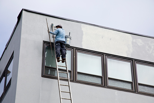 Elmwood Park, New Jersey – September 8, 2023: Industrial painter precariously high on unstable ladder working on office building.