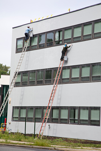 Elmwood Park, New Jersey – September 8, 2023: Industrial painters precariously high on unstable ladders working on office building.