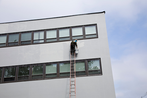 Elmwood Park, New Jersey – September 8, 2023: Industrial painter precariously high on unstable ladder working on office building.