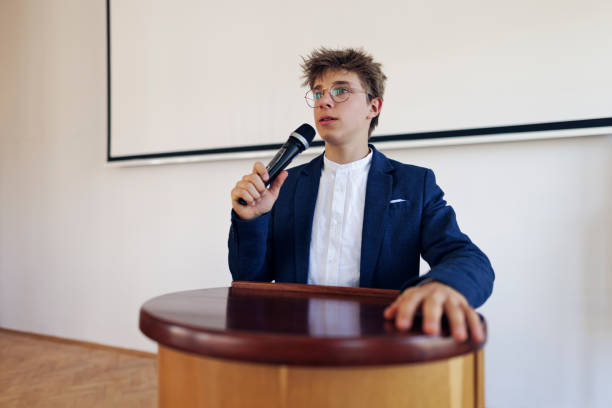 Teenage boy giving a speech on school assembly. The boy is holding a microphone.\nShot with Canon R5