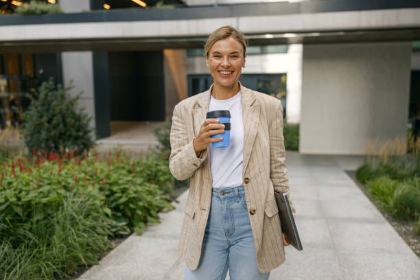 istock Business woman going to work while drinking coffee on office building background and looks camera 1670258405