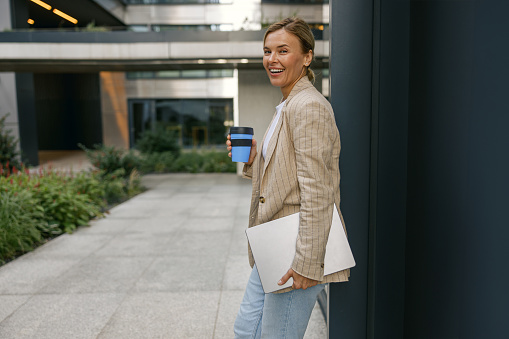 Female manager standing with laptop and take away coffee on stylish office yard and looks camera