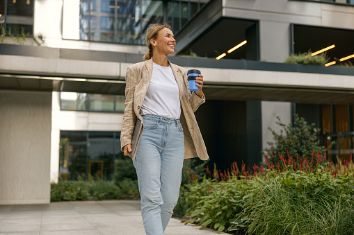 Stylish business woman drinking coffee during break time near office building and looks away