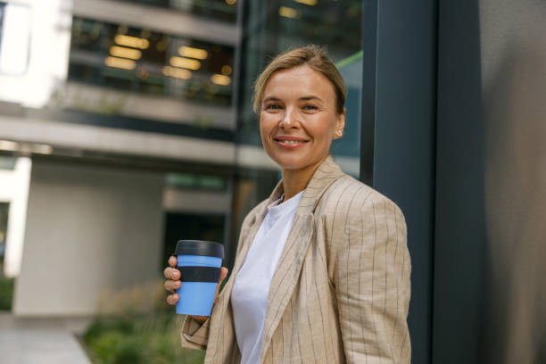 istock Business woman going to work while drinking coffee on office building background and looks camera 1670288151