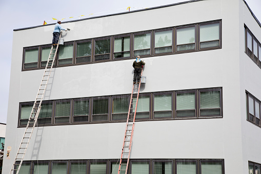 Elmwood Park, New Jersey – September 8, 2023: Industrial painters precariously high on unstable ladders working on office building.