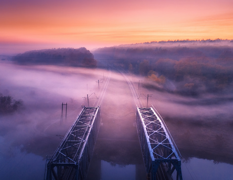 Aerial view of railroad bridge in fog at sunrise in autumn. Colorful landscape with foggy trees, fields, mist, river, railway station, red sky. Top view. Railroad in fog in dusk in fall. Industrial