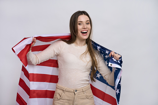 Beautiful patriotic vivacious young woman with the American flag held in her outstretched hands, standing isolated over white background, smiling looking at camera
