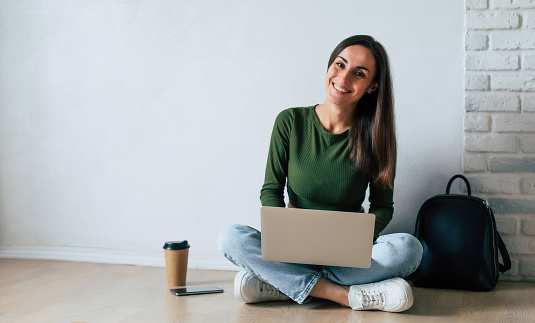 Young cute confident student woman in casual clothes is using a laptop and smiling while sitting on the floor, on gray background. Study, education, university, college, graduate concept