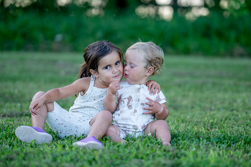 A sweet little three year old girl and her one year old brother, sit in the grass together on a sunny summers evening.  They are both dressed casually as they embrace and smile for a portrait.