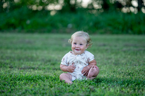 A sweet little one year old boy sits in the grass on a sunny summers evening.  He is dressed casually and smiling as he poses for a portrait.