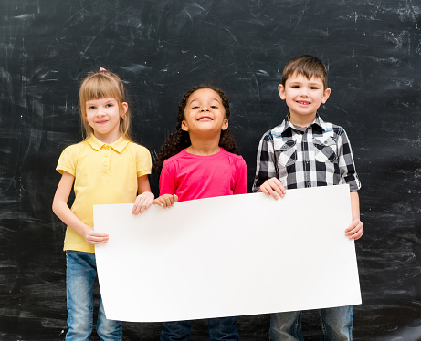 three cute children holding an empty paper sheet for ad with chalky blackboard on background