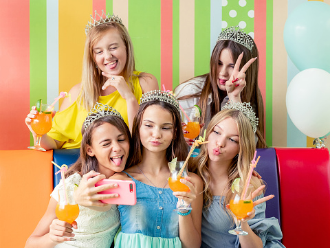 Pretty smiling teenage girls in dresses and crowns sit hugging together holding beverages and taking selfie at birthday party on the striped colorful background