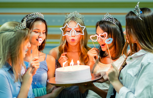Pretty smiling teenage girls in dresses and crowns holding cake at birthday party od