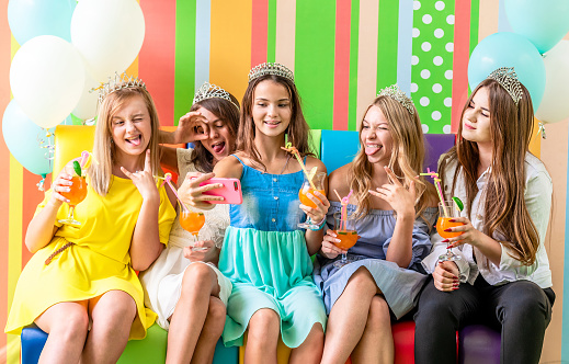 Pretty smiling teenage girls in dresses and crowns sit hugging together holding beverages and taking selfie at birthday party on the striped colorful background