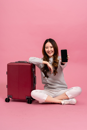 Beautiful asian female passenger showing her smart phone. Portrait of a smiling girl in sweater with suitcase in full length size. Winter lifestyle and travel concept