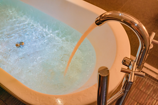 In soft lighting, a faucet is running hot water into a bathtub, preparing for a bath.