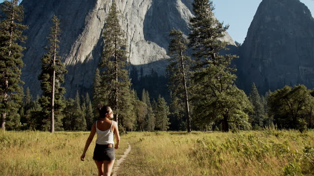 Young attractive woman walking trail in meadow looking up at rock face in Yosemite National Park